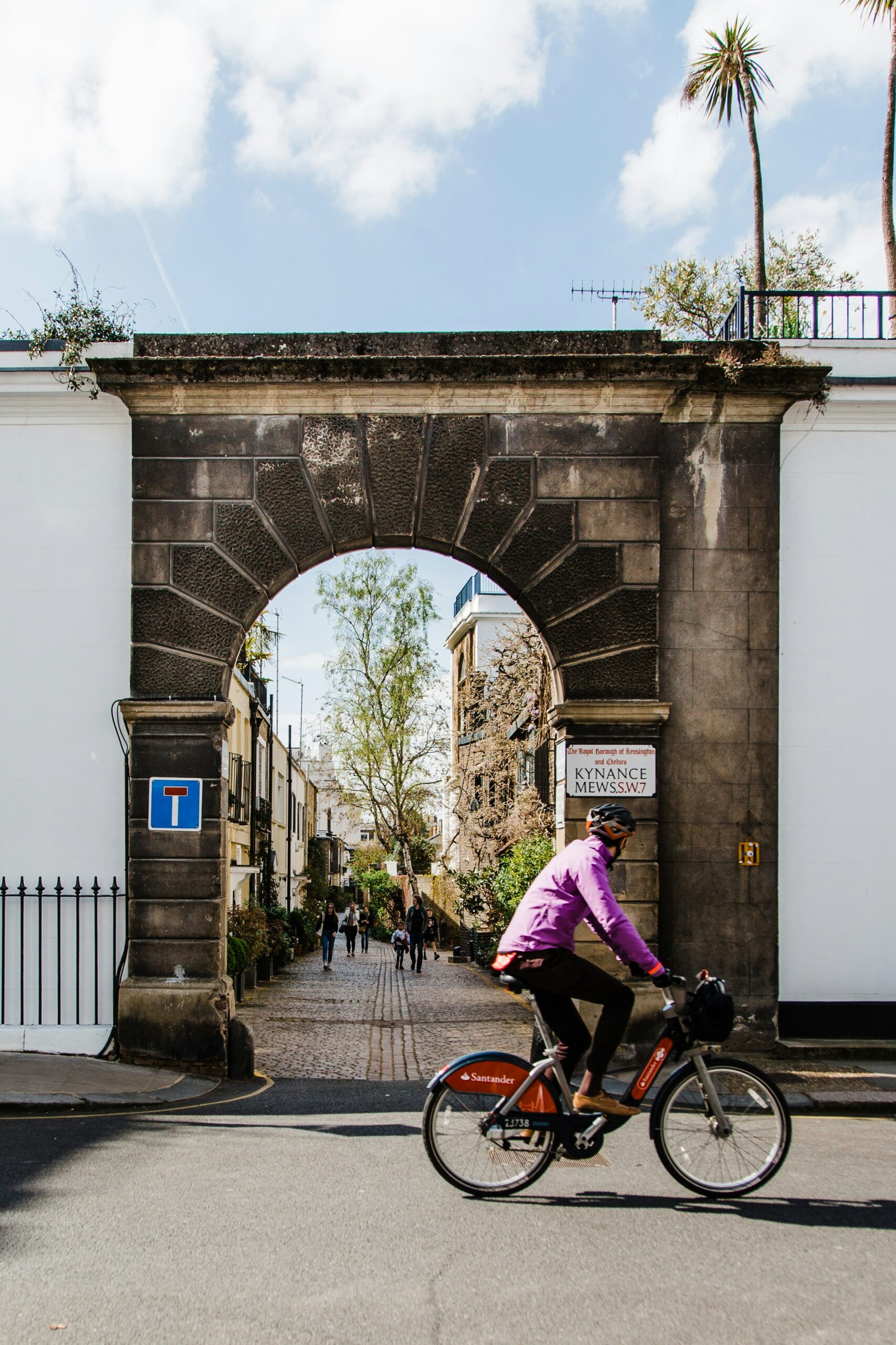 Kynance Mews, London – A picturesque cobbled mews street in London, framed by a historic stone archway with a cyclist riding past on a Santander rental bike. The charming alleyway, lined with ivy-covered houses, exudes a classic London charm, perfect for photography.