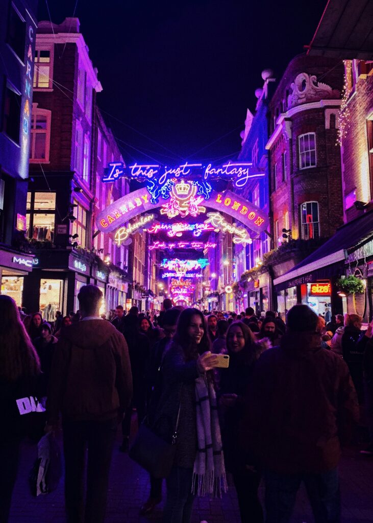 Carnaby Street at Night, London – A lively night scene on Carnaby Street, illuminated by vibrant neon signs inspired by Bohemian Rhapsody, including the phrase "Is this just fantasy?" A crowd of people strolls through the bustling shopping district, with two women in the foreground taking a selfie under the colourful lights