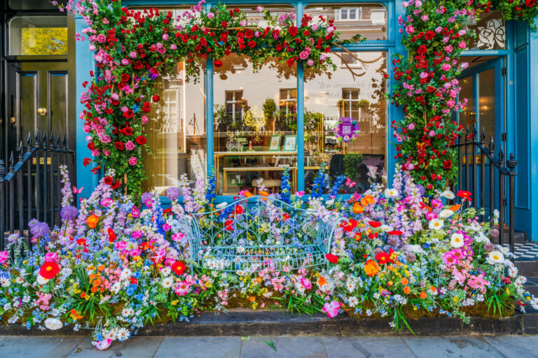 A storefront extravagantly decorated for Chelsea in Bloom, with a vibrant explosion of flowers in front of and around the window. The display includes a pastel-blue wrought-iron bench surrounded by colorful blooms like daisies, poppies, delphiniums, roses, and alliums in shades of red, pink, orange, purple, and white, creating a whimsical garden effect.